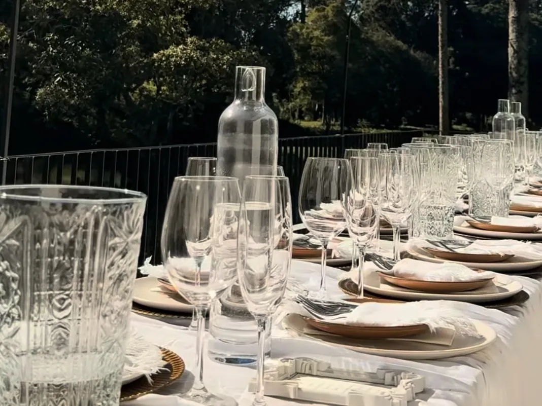 Mesa longa de evento corporativo posta ao ar livre, com toalha branca, taças e pratos, em meio às araucárias da propriedade.