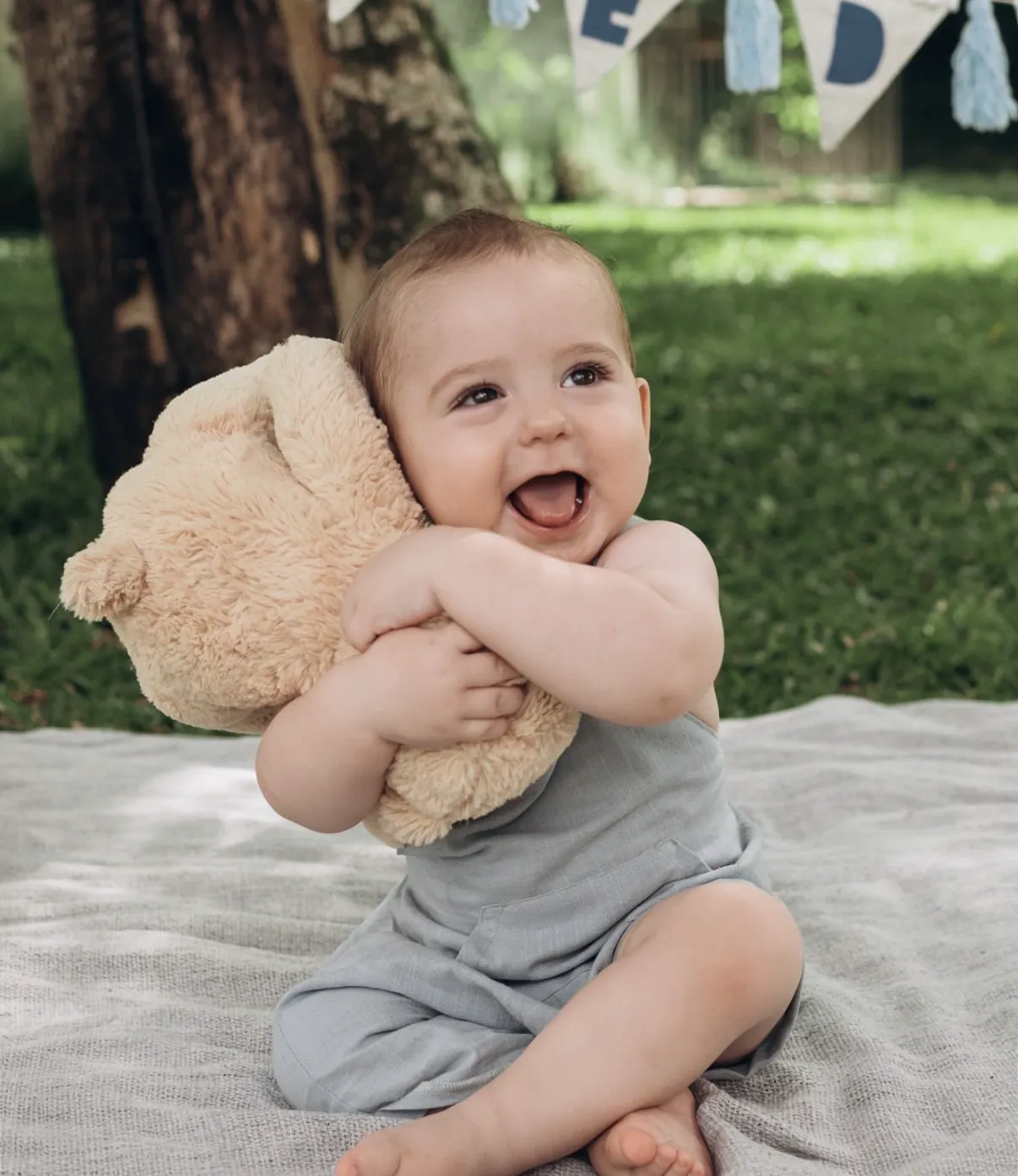 Bebê sorrindo ao ar livre abraçado a um ursinho de pelúcia, sentado sobre uma manta clara no gramado com bandeirinhas ao fundo.