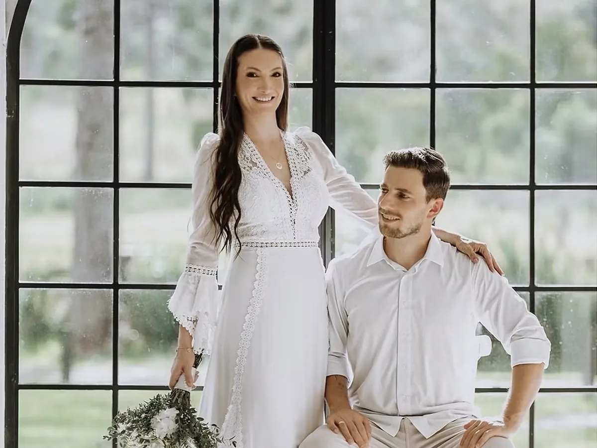 Casal em ensaio pré-wedding: noiva sorridente de vestido branco de renda com buquê, em pé, e noivo de camisa branca sentado ao lado, diante da grande janela arqueada de vidro com as araucárias ao fundo.