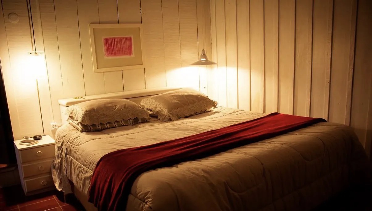 Quarto da Casa de Vidro à noite, com cama de casal de edredom bege, manta vermelha, travesseiros de renda e luz quente de abajur sobre a parede de madeira.