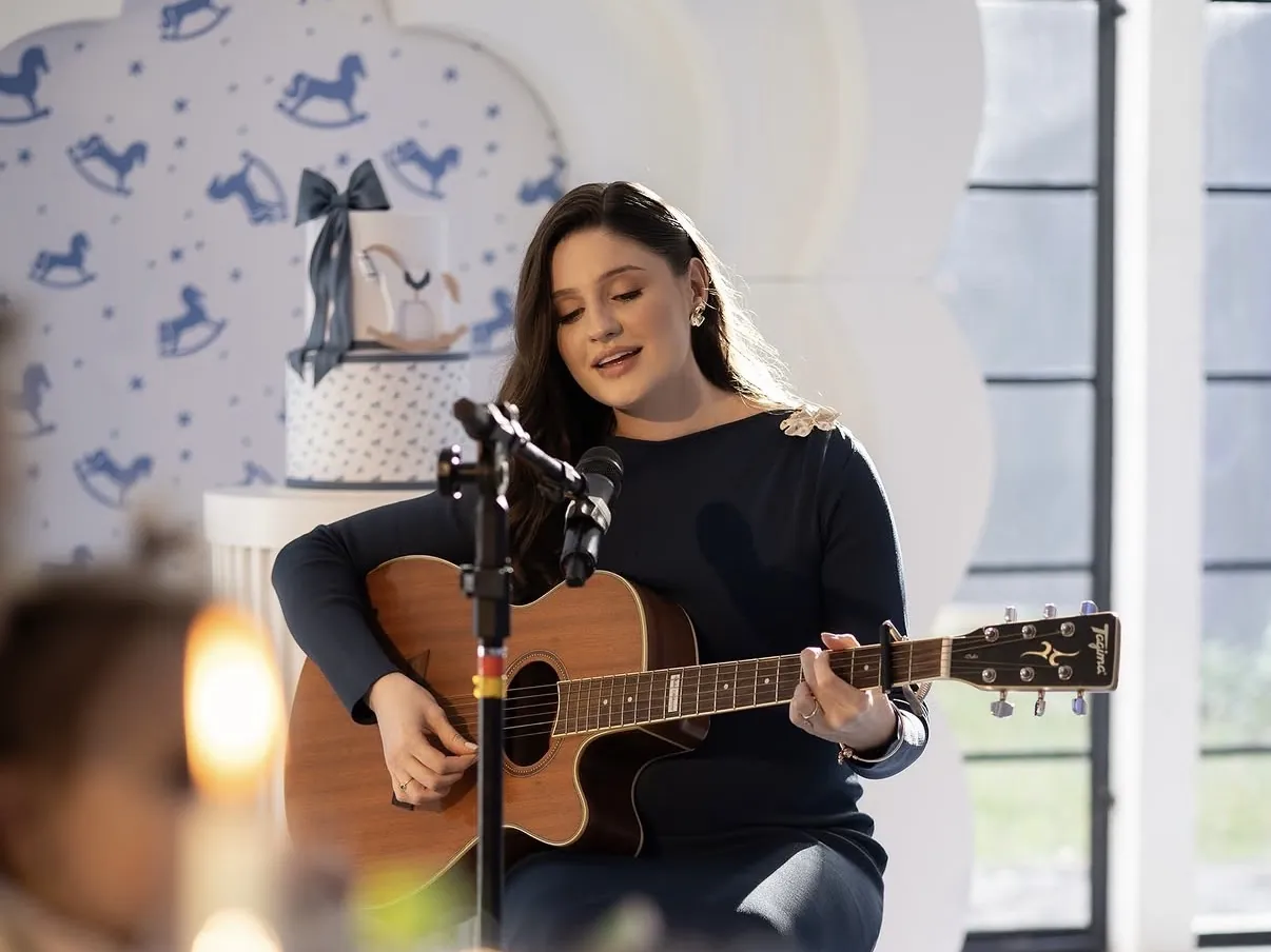 Cantora ao violão se apresentando em festa no salão, com bolo decorado e mesa posta em primeiro plano.