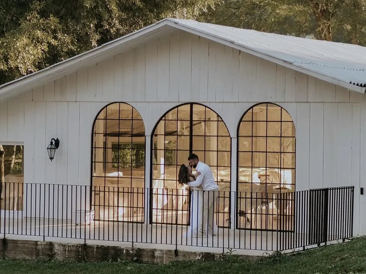 Casal se beijando na varanda do salão de fachada branca com portas em arco, com o gramado e as árvores da propriedade ao redor.