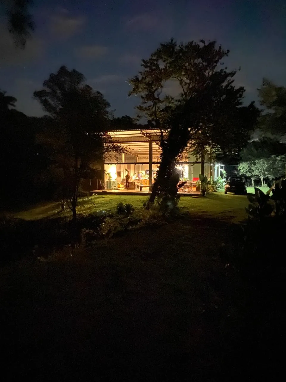 Casa de Vidro à noite vista do gramado, com as paredes de vidro iluminadas por dentro e o jardim escuro em volta.