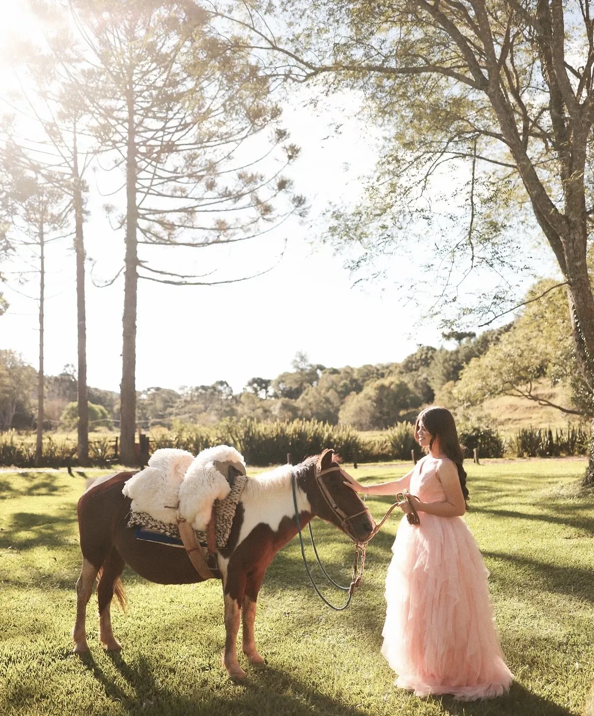 Moça de vestido rosa acariciando um cavalo com sela branca no gramado do sítio, com araucárias ao fundo.