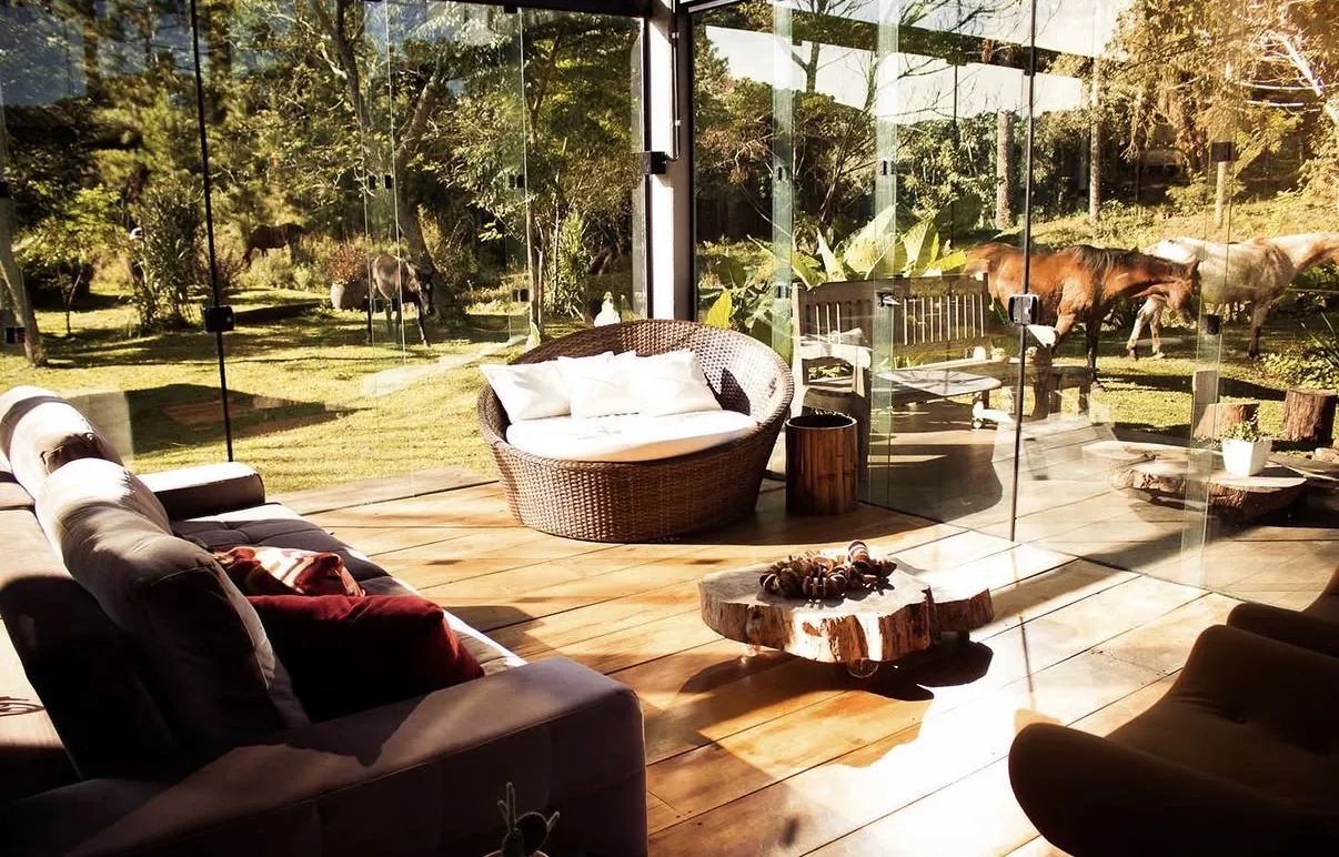 Sala de estar da Casa de Vidro com sofás e poltronas diante das paredes de vidro, com cavalos pastando no gramado lá fora.