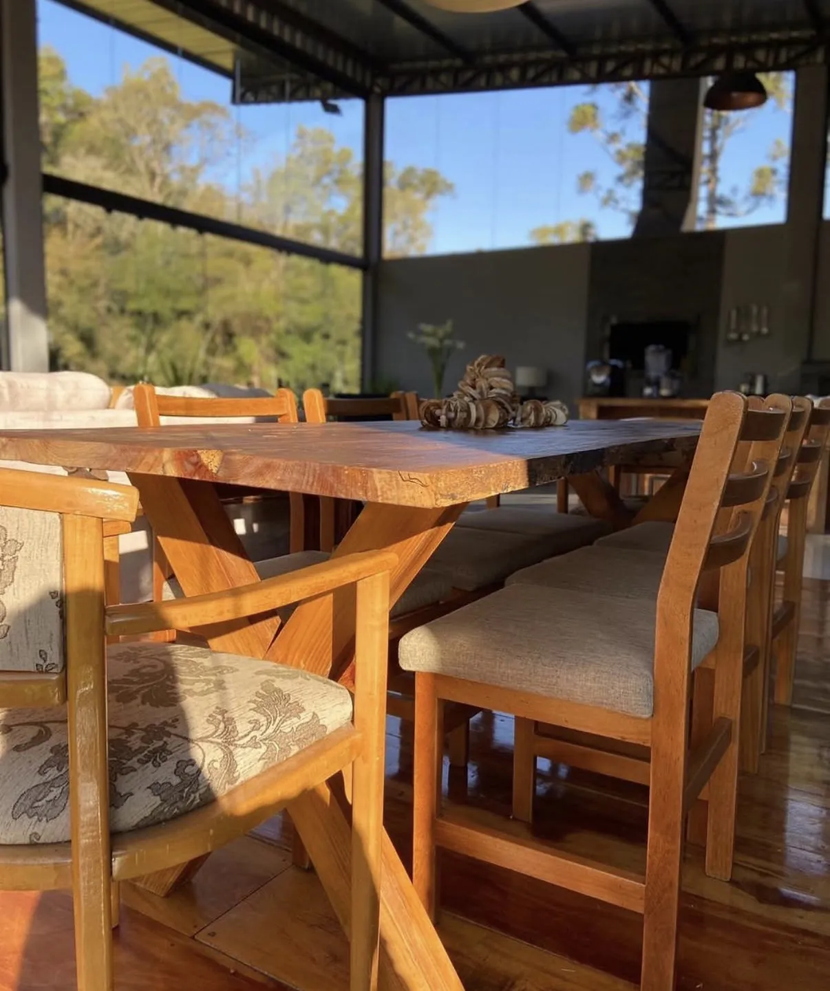 Mesa de jantar de madeira maciça com cadeiras estofadas e cozinha ao fundo, com a natureza vista pelas paredes de vidro da Casa de Vidro.